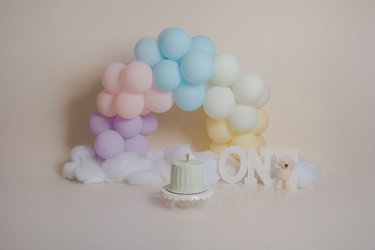 Pastel balloon arch with white cake stand and 'ONE' letters for birthday celebration.