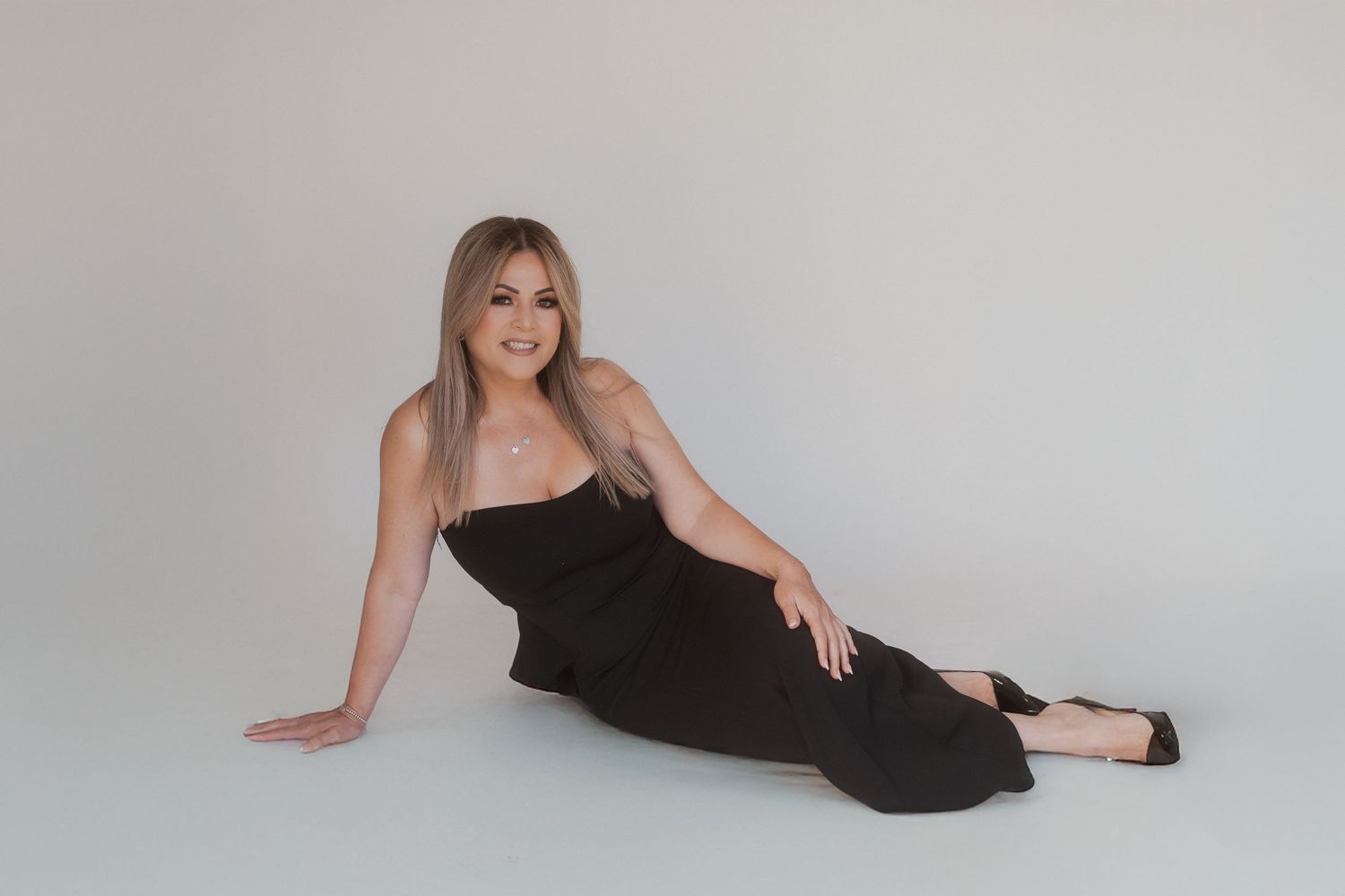 Woman in elegant black dress poses gracefully on studio floor against light grey background.