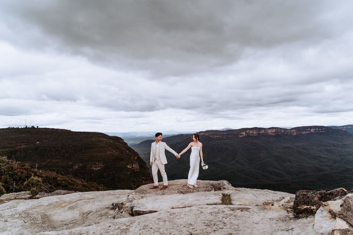 Couple holding hands on mountain cliff overlooking scenic valley.