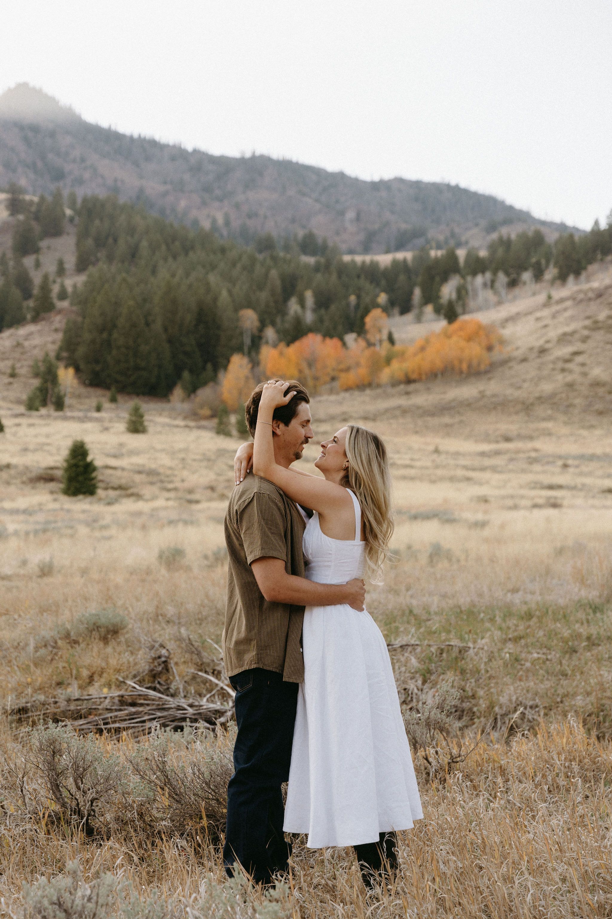 Two people embrace in a romantic moment against an autumn mountain landscape with golden trees and rolling hills.