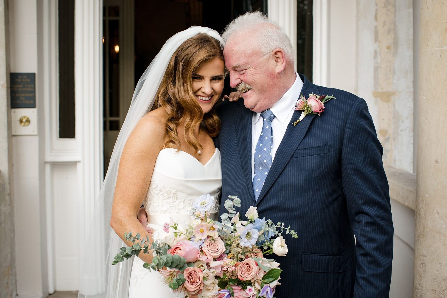 A bride in a white dress holds a pink and blue floral bouquet while sharing a joyful moment at a wedding venue in Halifax, Nova Scotia.