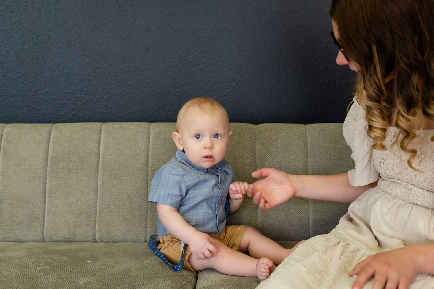 Baby in gray t-shirt sits on gray couch while someone offers them something from the side.