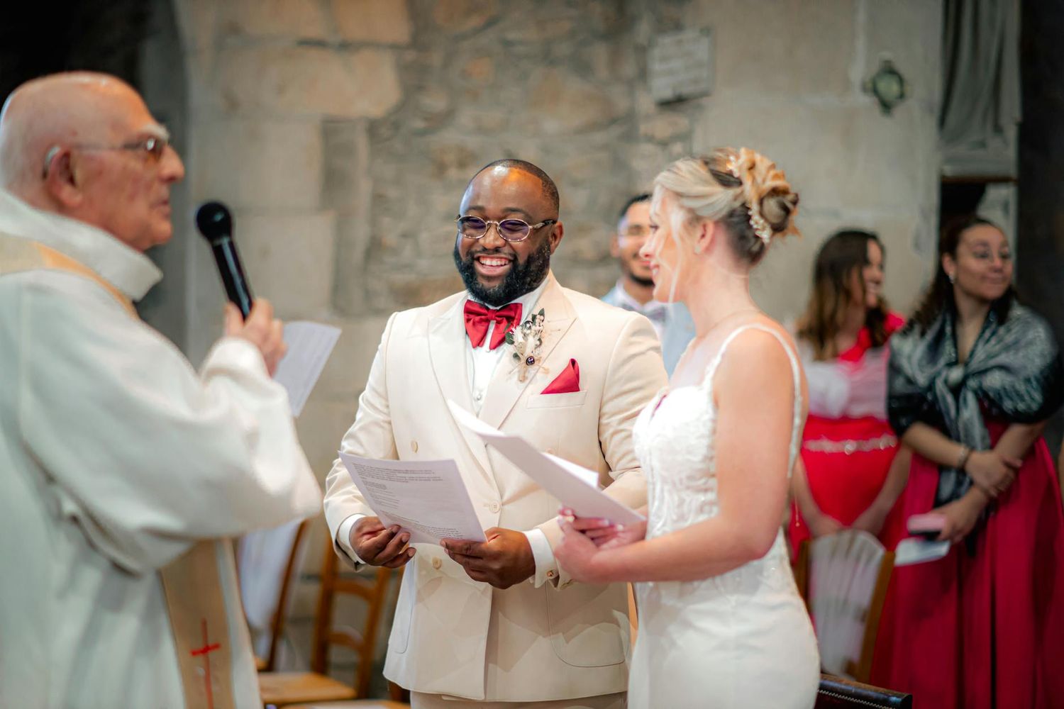 A couple stands with an officiant during their wedding ceremony, with guests watching in the background.