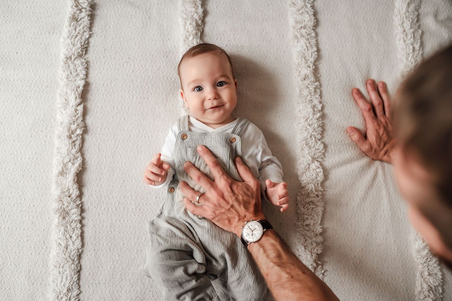 A smiling baby wearing gray clothing lies on a textured blanket while adult hands reach into the frame.