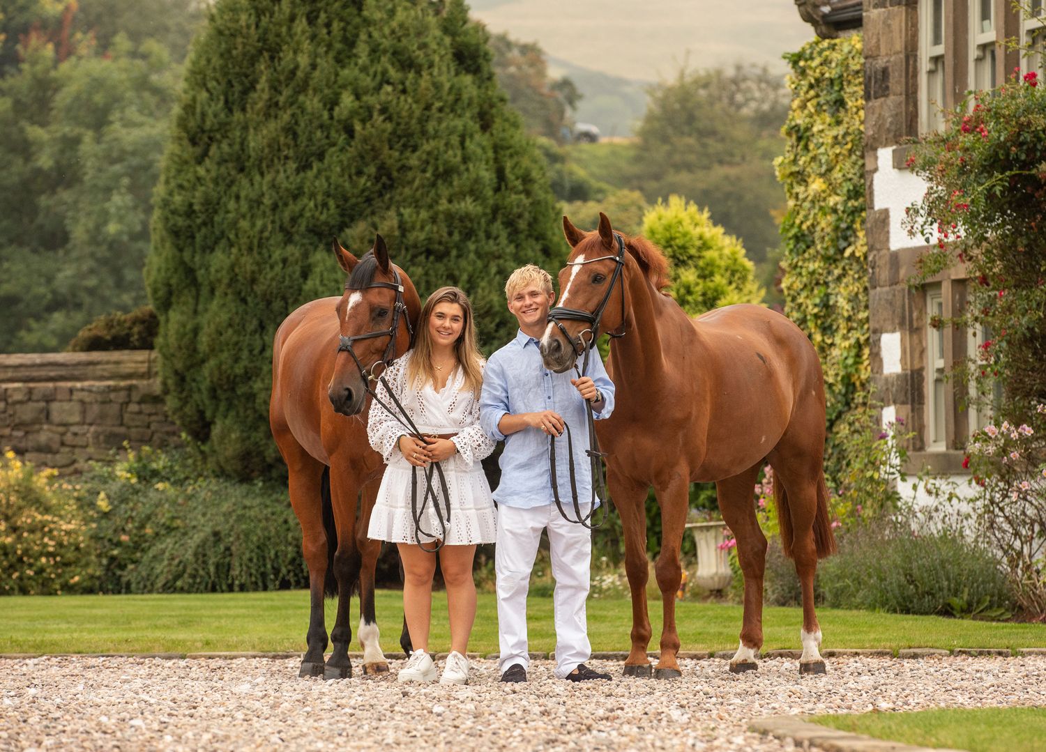 Two horses and their companions stand on a gravel driveway in front of a stone building.