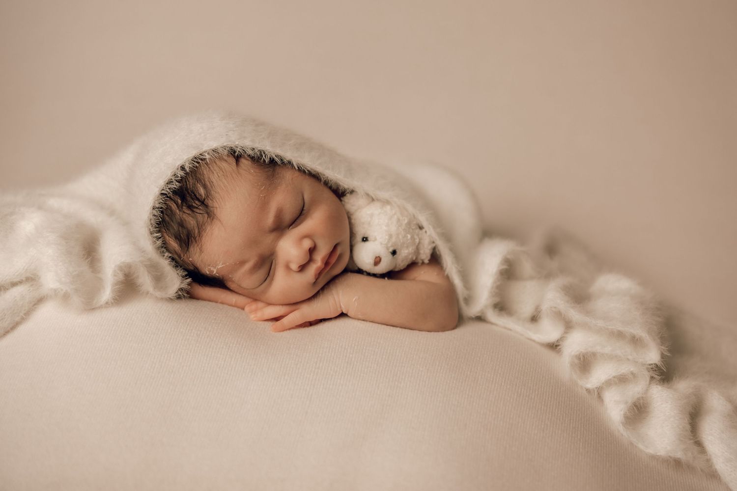 A sleeping newborn wrapped in soft white fabric on a beige backdrop.