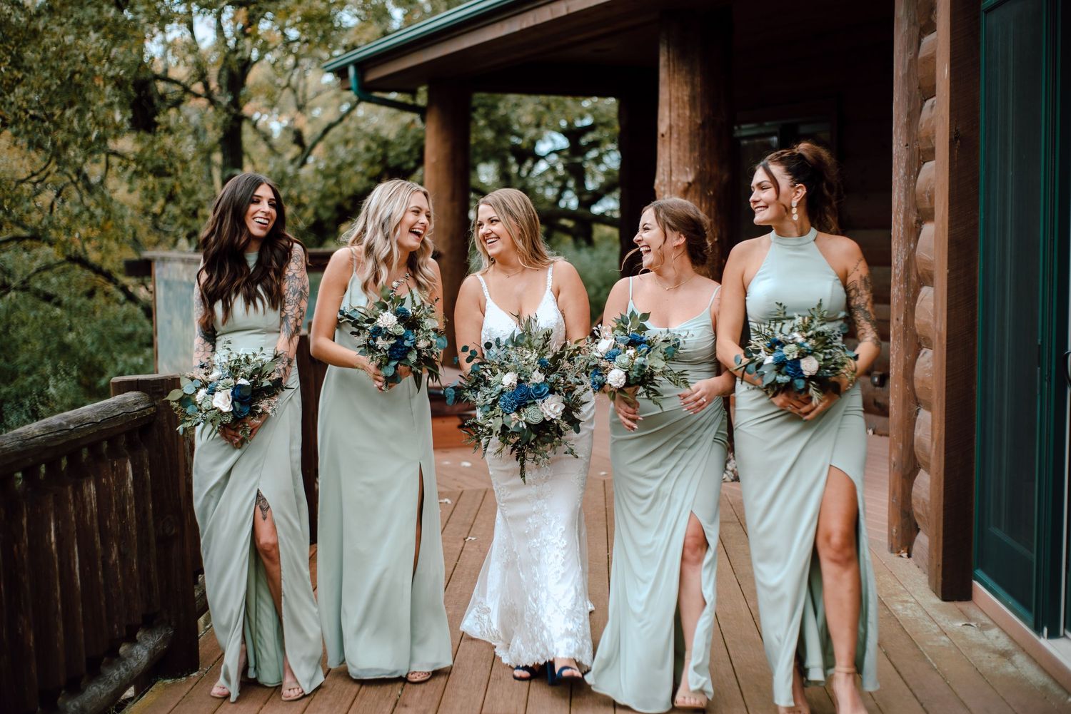 Bridesmaids in matching sage green dresses pose with bouquets on a rustic wooden deck.