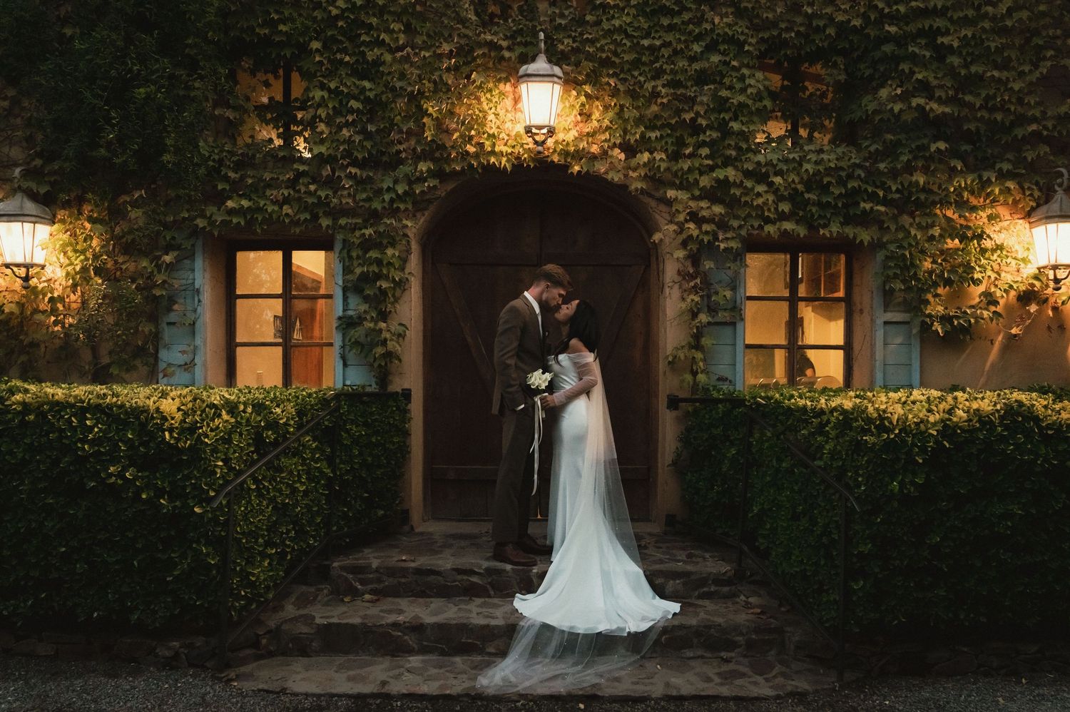 A romantic evening wedding scene with ivy-covered walls, glowing lanterns, and a couple embracing under a stone archway.
