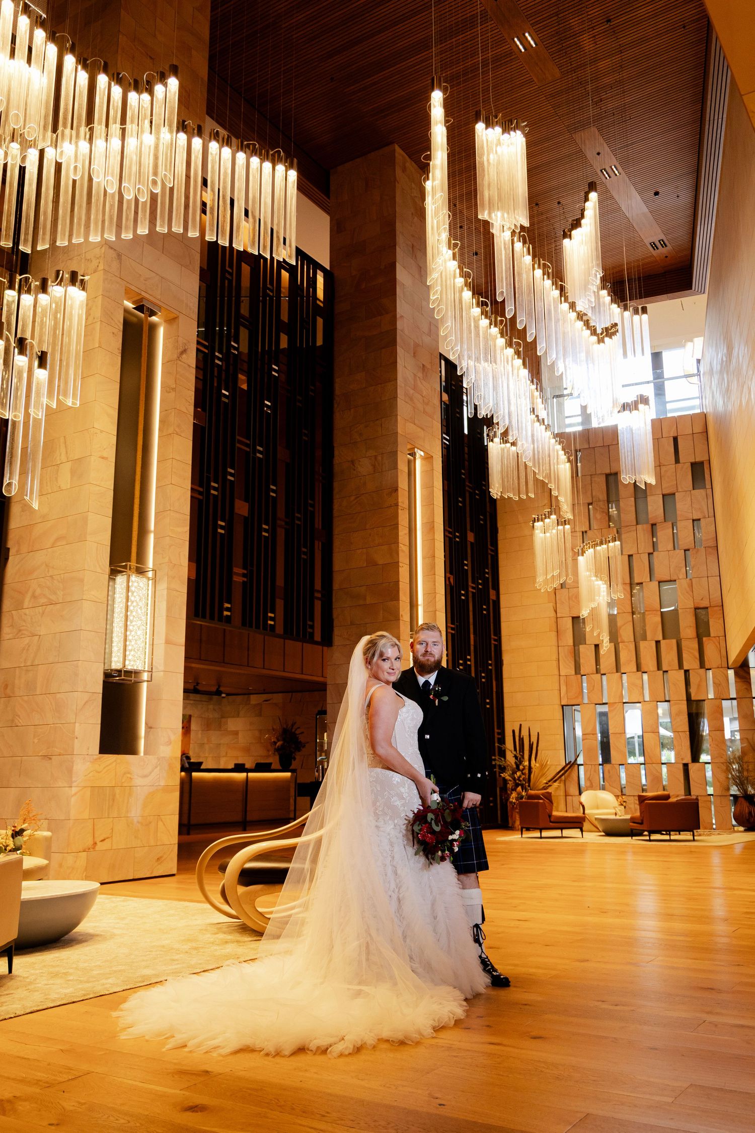 A newlywed couple shares a romantic moment in a grand ballroom with glowing vertical light fixtures illuminating the space.