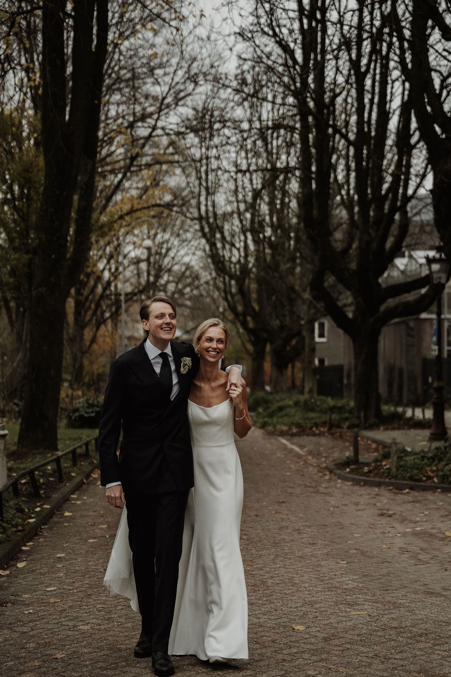 A couple walks together on a tree-lined path during their autumn wedding celebration.