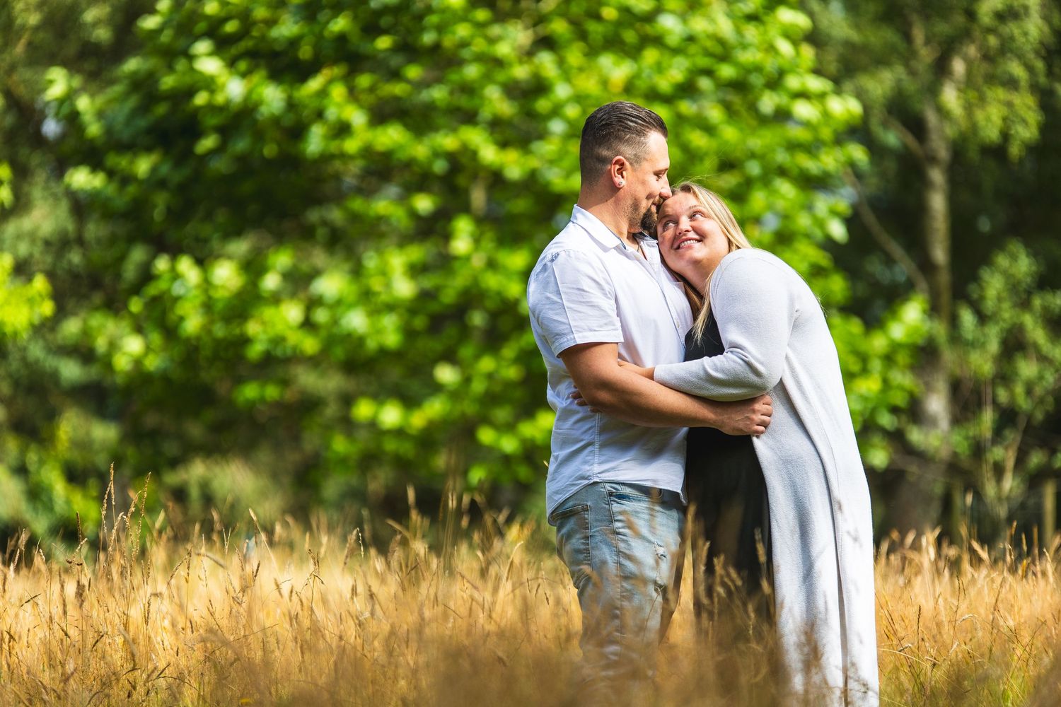 Couple sharing an intimate moment in a sunlit meadow with green trees in background.