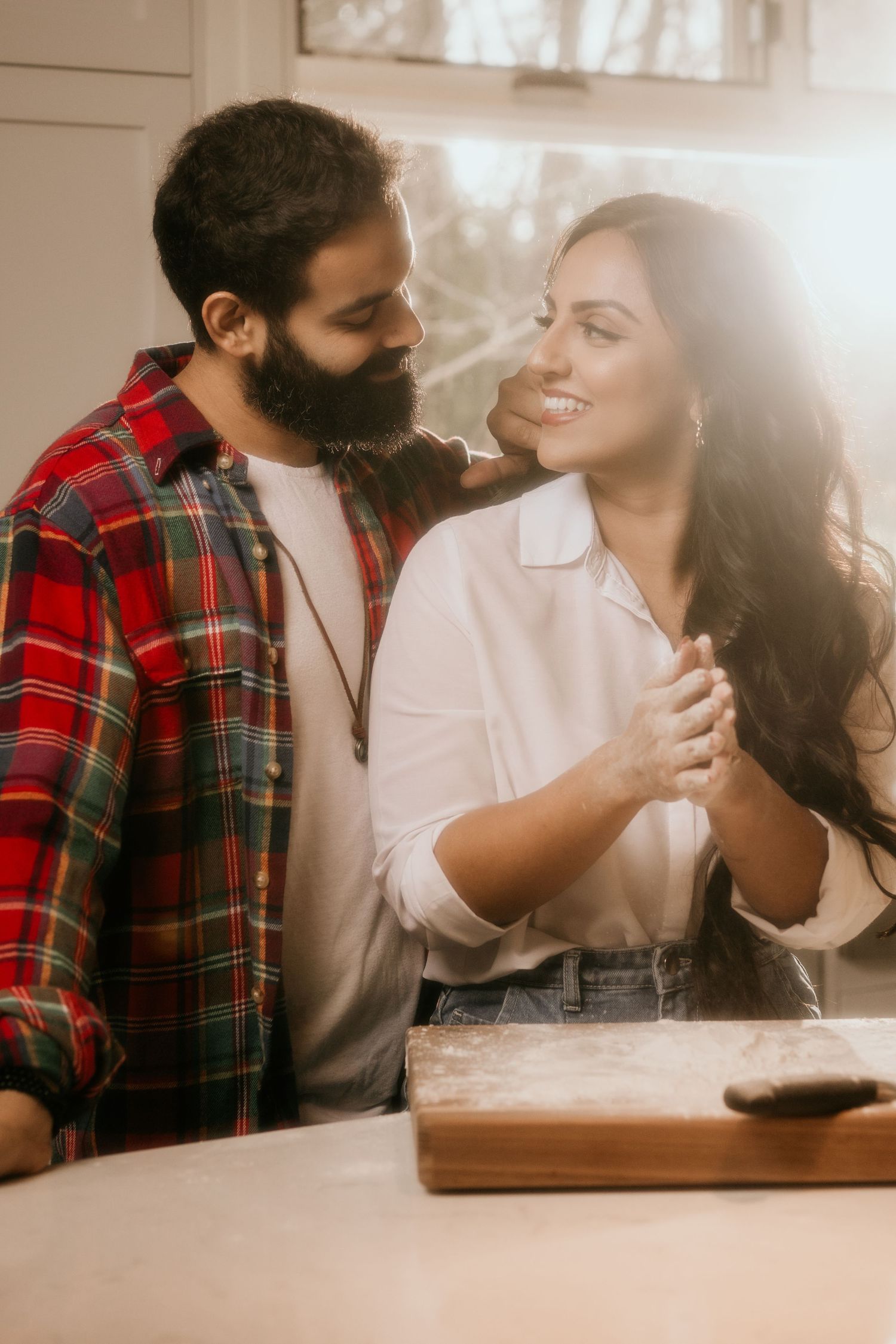 A couple shares an intimate moment in the kitchen while standing near a wooden cutting board in warm lighting.