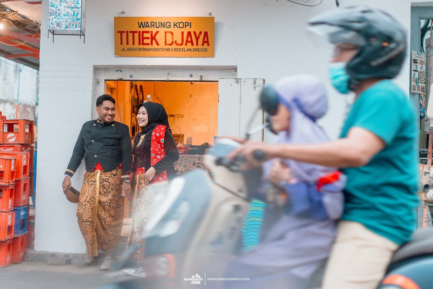 People exit a storefront with orange signage while a scooter rider passes by on a busy street in Indonesia.