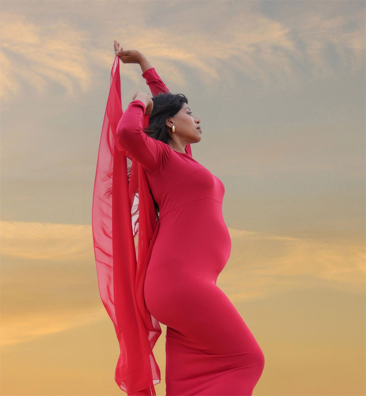 A pregnant figure in a flowing red dress poses gracefully against a sunset sky background during a maternity photoshoot.