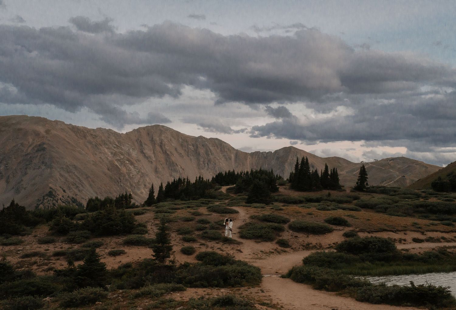 Two people walk across a rural landscape with rolling hills and dramatic clouds at sunset.