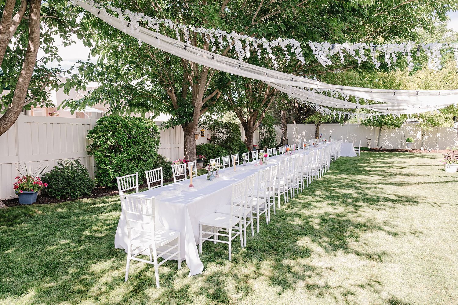 Long white banquet tables with chairs set up outdoors under string lights and trees for an elegant garden party setting.