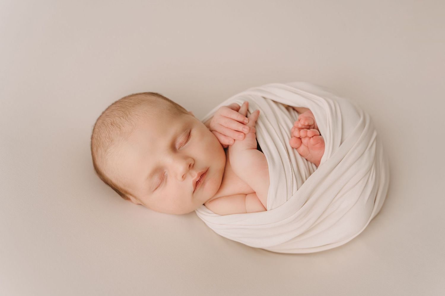 A peacefully sleeping newborn swaddled in soft white fabric against a neutral beige background.