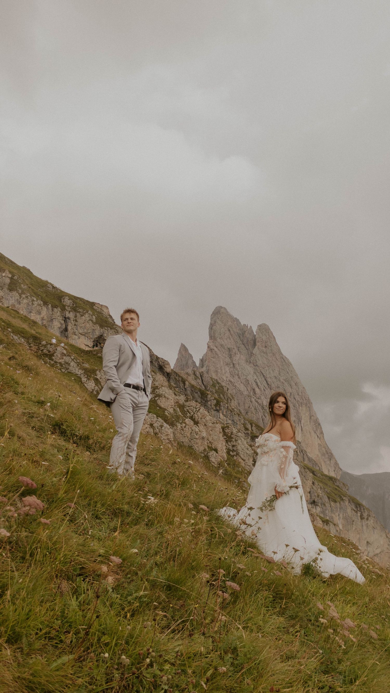 A couple in wedding attire poses on a mountainside with dramatic rocky peaks and cloudy skies in the background.