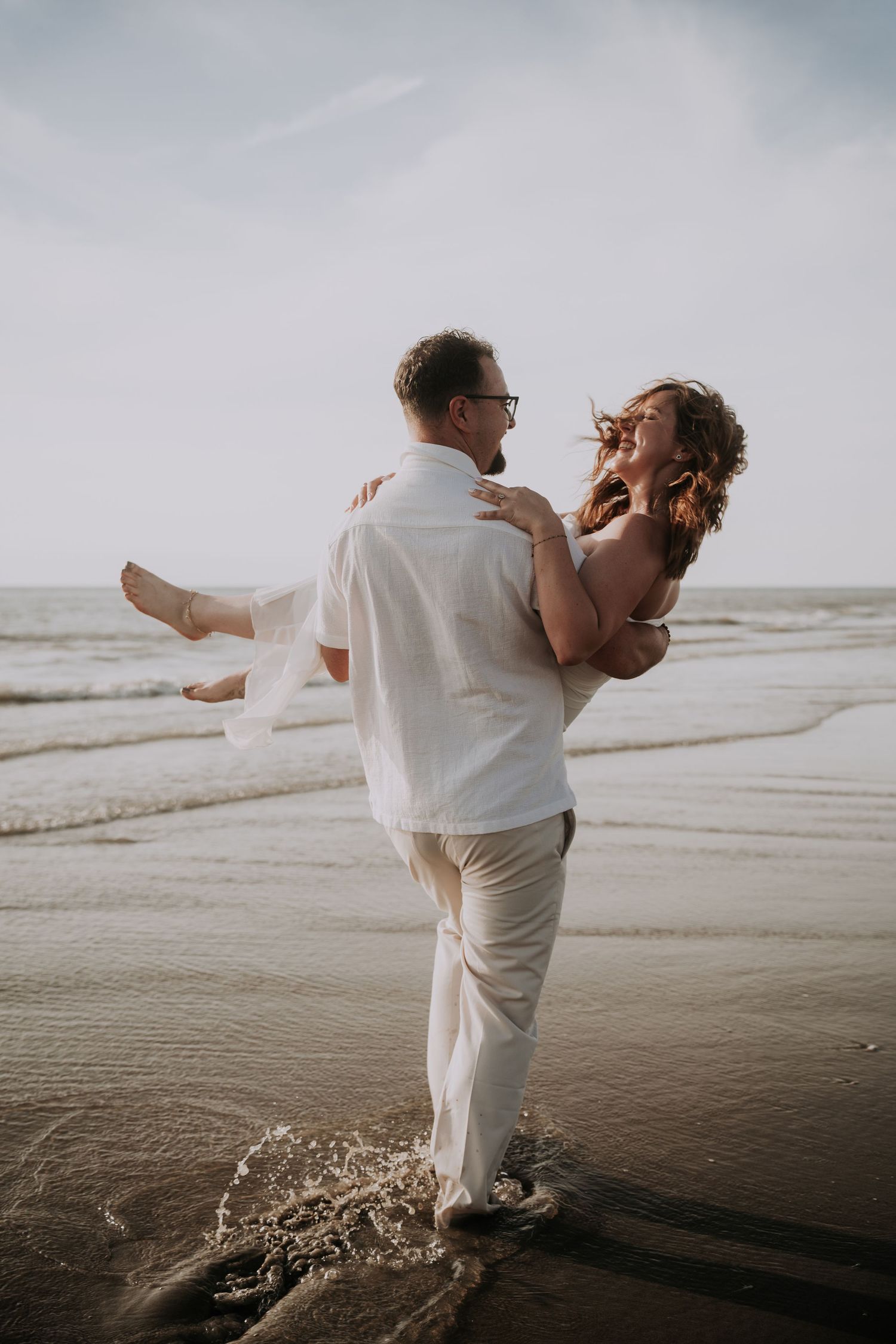 A couple shares a joyful moment on the beach, man carrying woman in white outfits with ocean waves behind them.