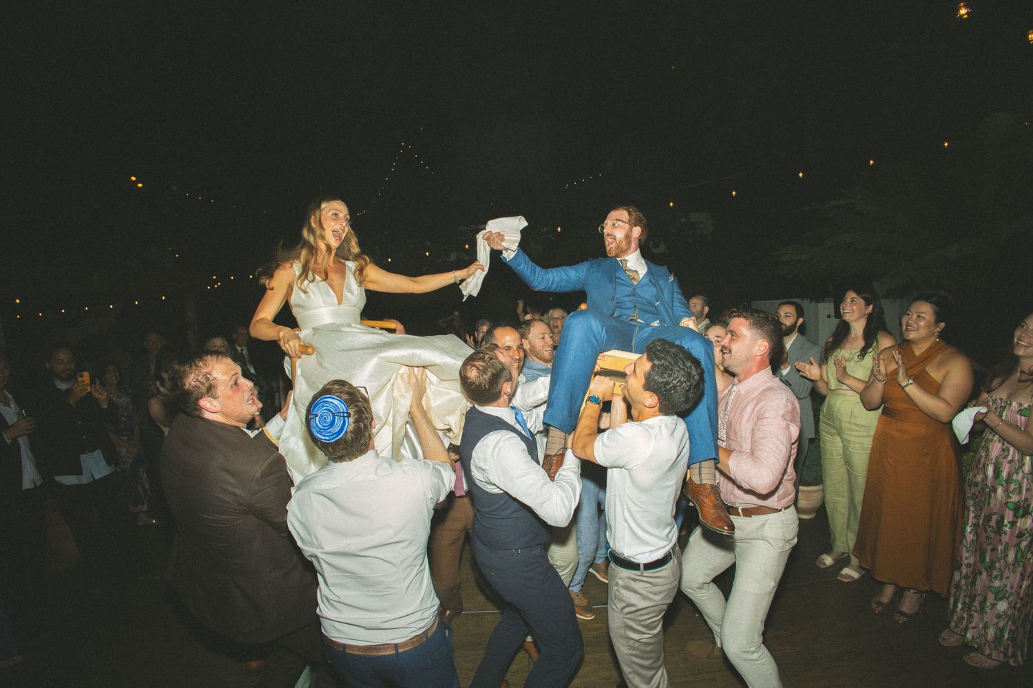 Bride and groom lifted on chairs during joyful Jewish wedding hora dance celebration surrounded by cheering guests at night.