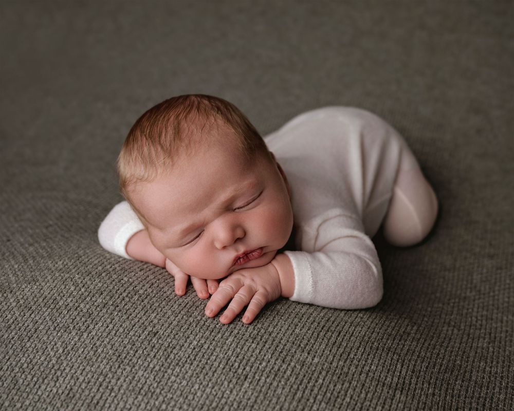 Peaceful newborn baby sleeping in fetal position on gray background wearing white onesie in professional studio photo.