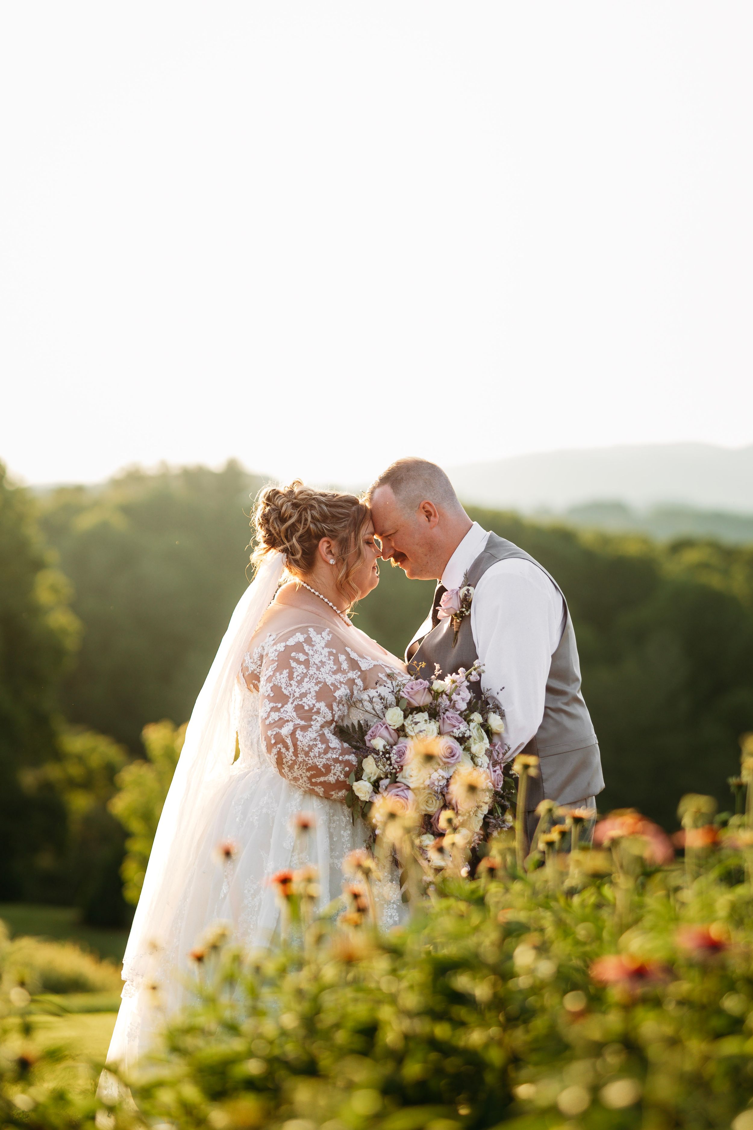 Bride and groom touching foreheads in golden hour light, surrounded by blooming flowers with rolling green hills behind them.