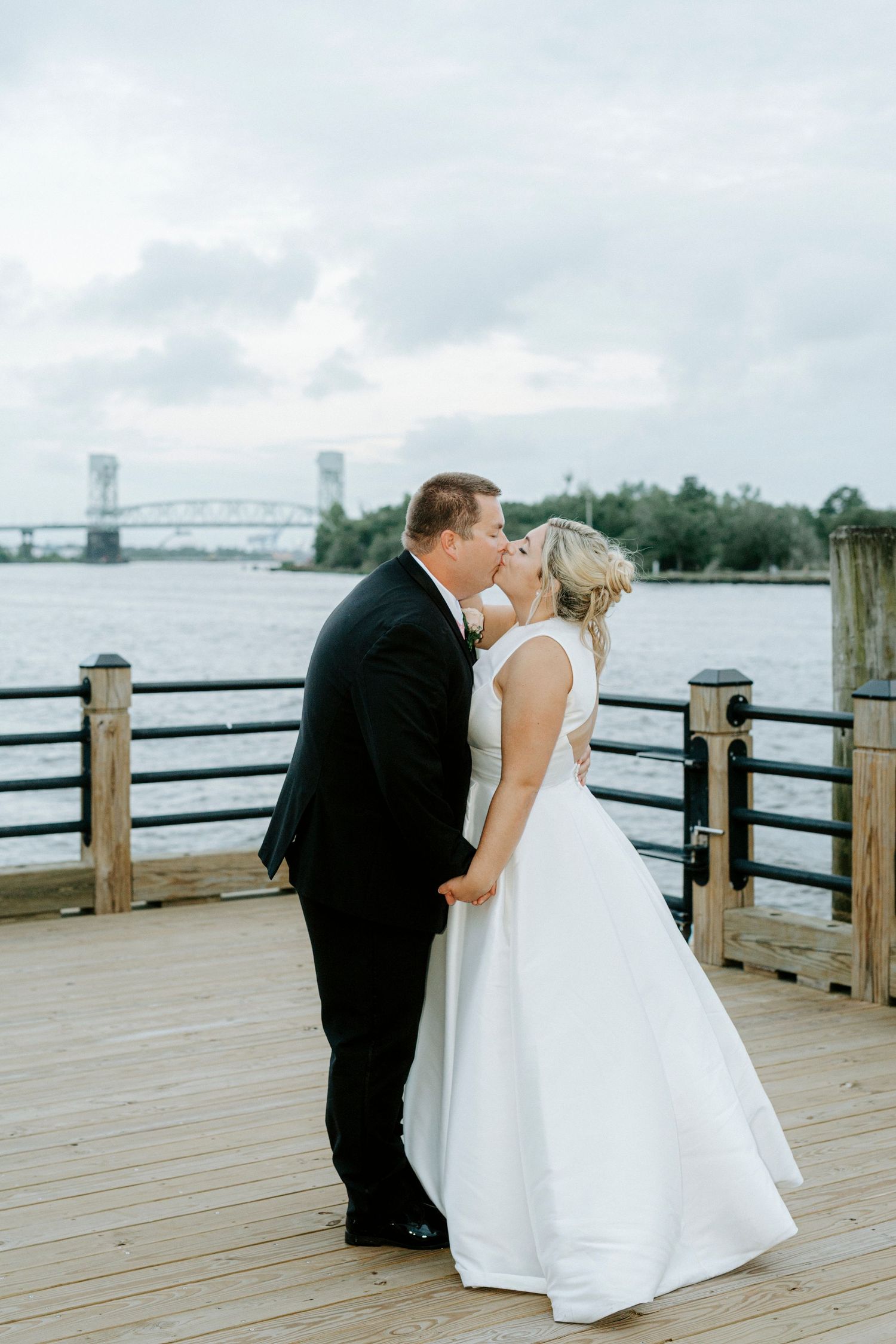 Bride and groom share a kiss on a waterfront dock with a bridge and river in the background on their wedding day.