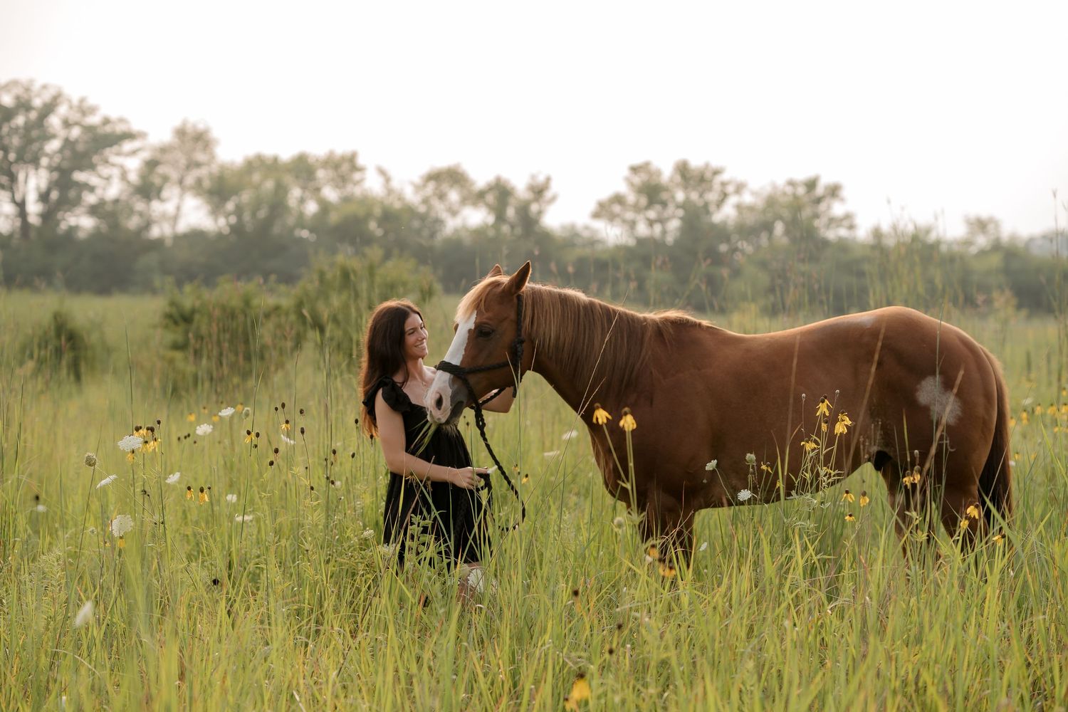 Young woman in a field bonding with a chestnut horse among wildflowers with trees in the background.