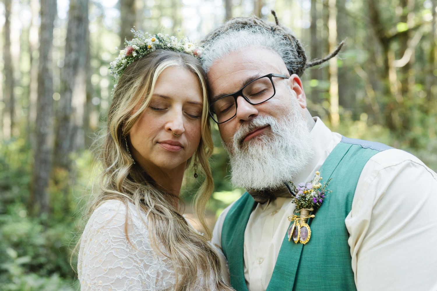 A bride and groom share a tender moment in a forest, she wearing a floral crown and he a green vest with boutonniere.
