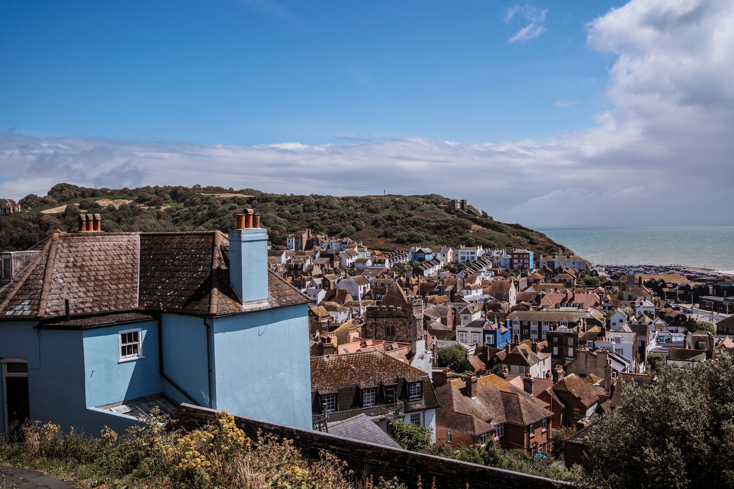 Aerial view of a charming coastal town with colorful rooftops, lush green hills, and a blue sea under a partly cloudy sky.