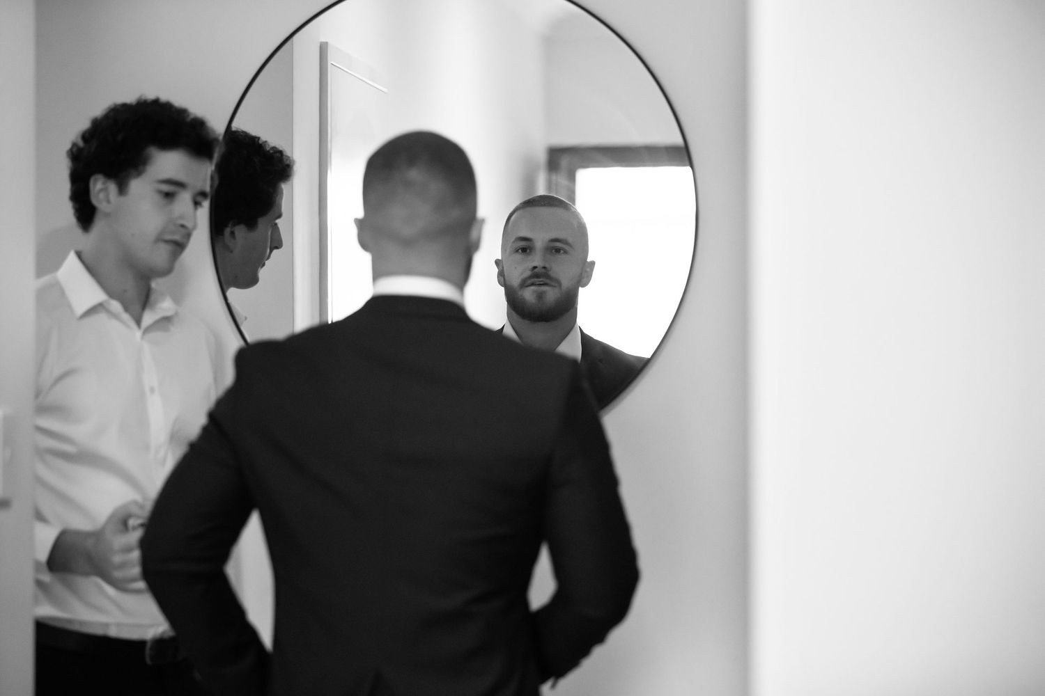 Two men in formal attire stand before a round mirror in a black and white photo.