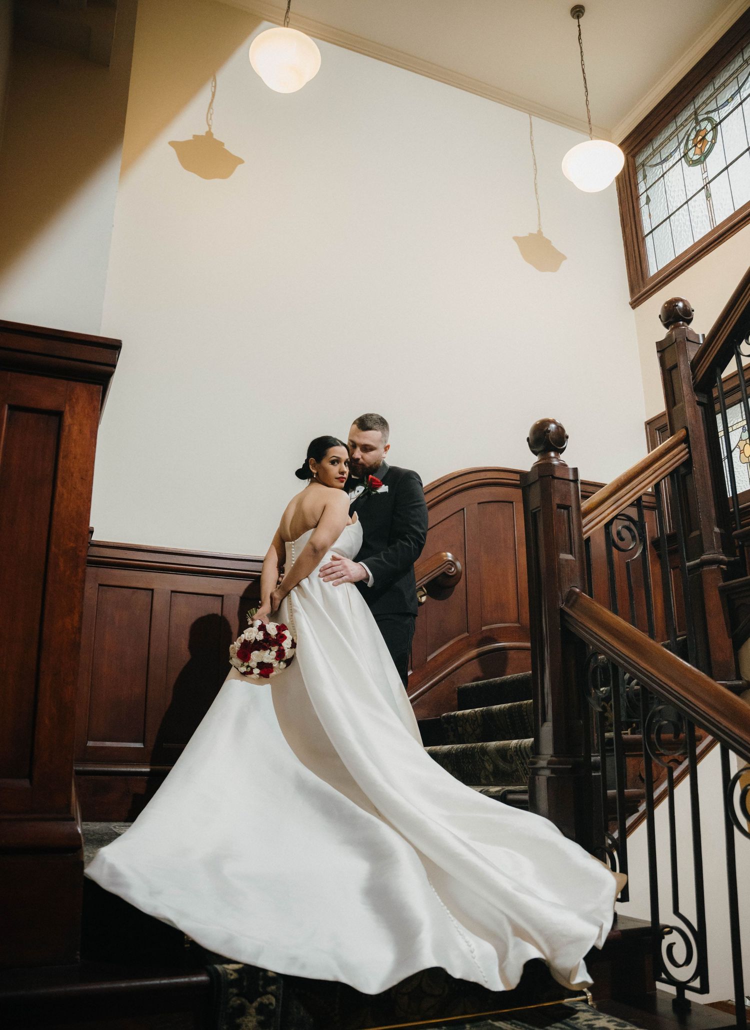 Bride and groom embrace on grand wooden staircase, her white gown flowing, in an elegant historic venue.