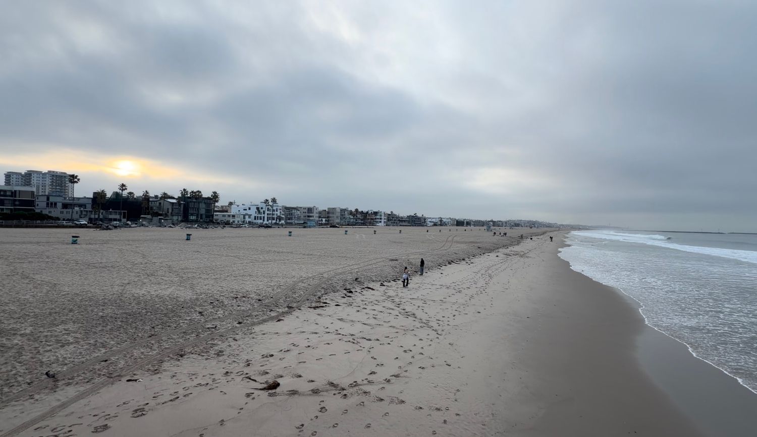 Overcast beach shoreline with wet sand, gentle waves, and distant buildings under a cloudy sky at dusk.