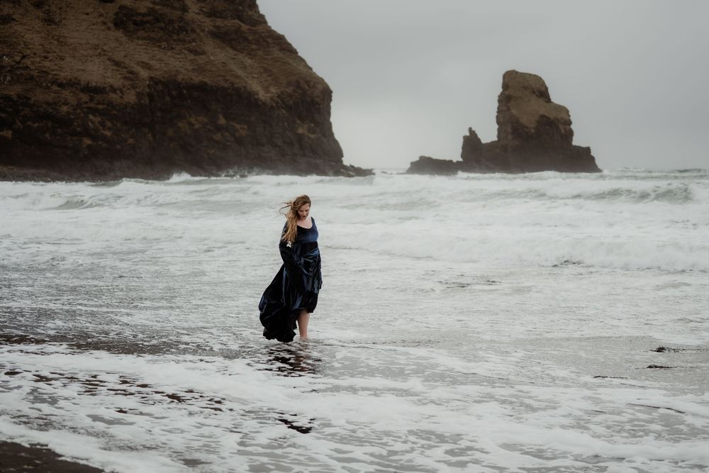 Woman in dark dress walking along a moody, wave-washed beach with rocky cliffs in the background.