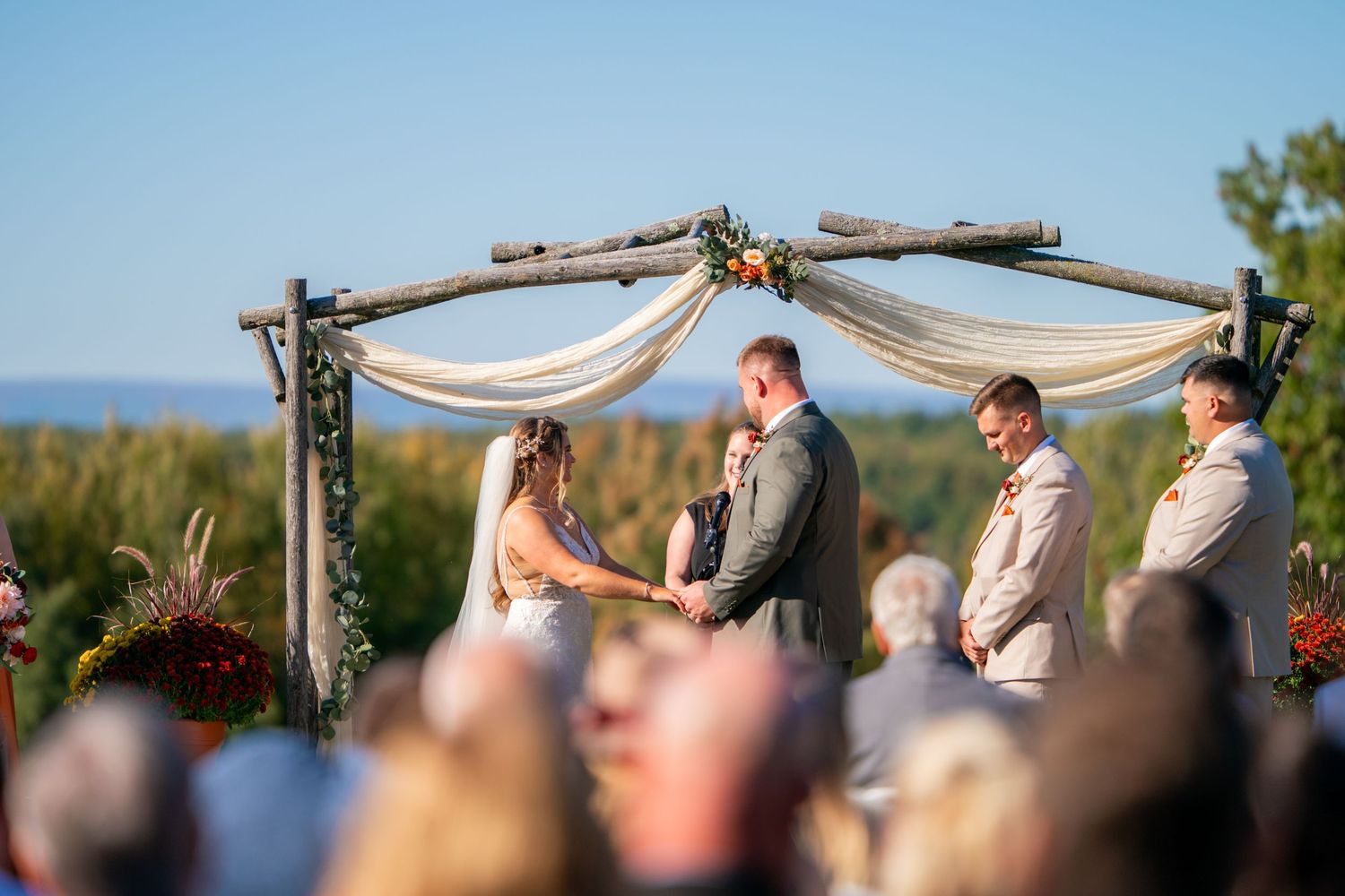 A wedding ceremony takes place outdoors under a wooden arch decorated with white fabric and flowers on a sunny day.