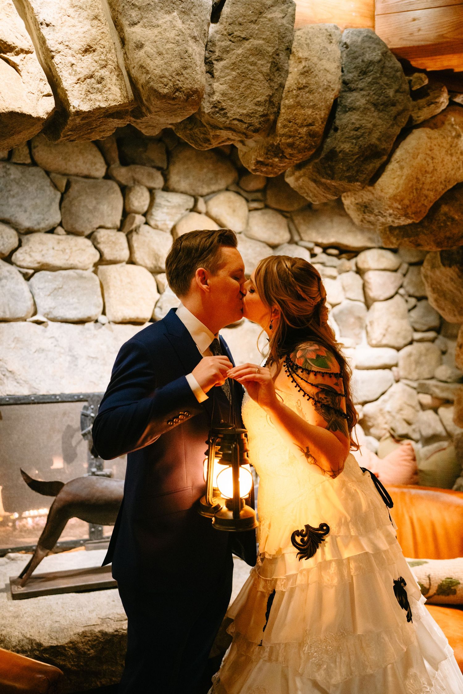 A romantic couple in formal wedding attire share an intimate moment against a rustic stone wall backdrop.