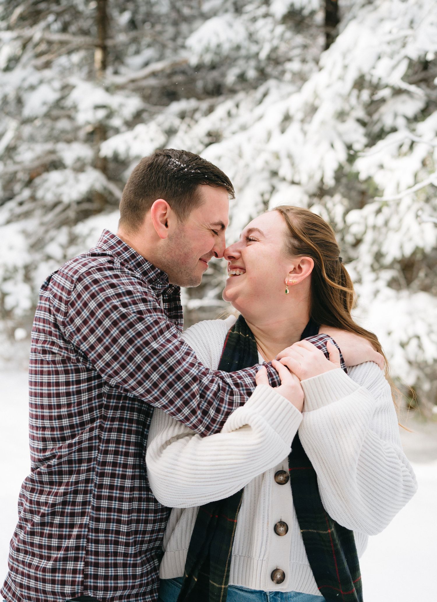 A couple shares a joyful moment together in a snowy winter forest setting with snow-covered trees in the background.
