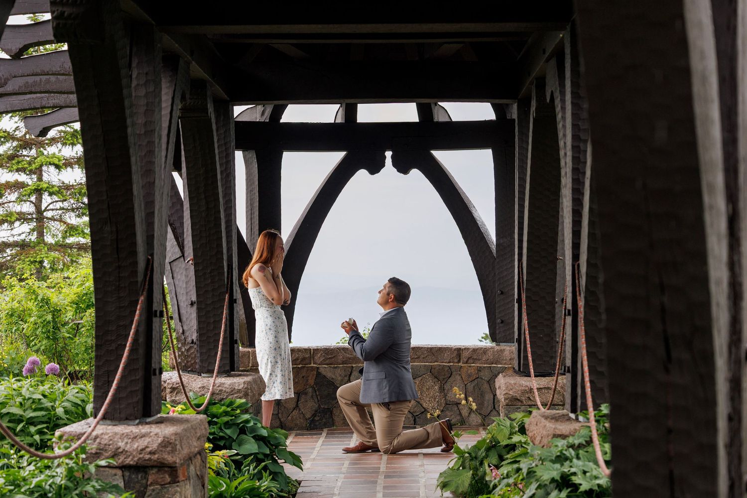 Man proposes on one knee to woman under ornate wooden pergola surrounded by lush greenery with ocean view.