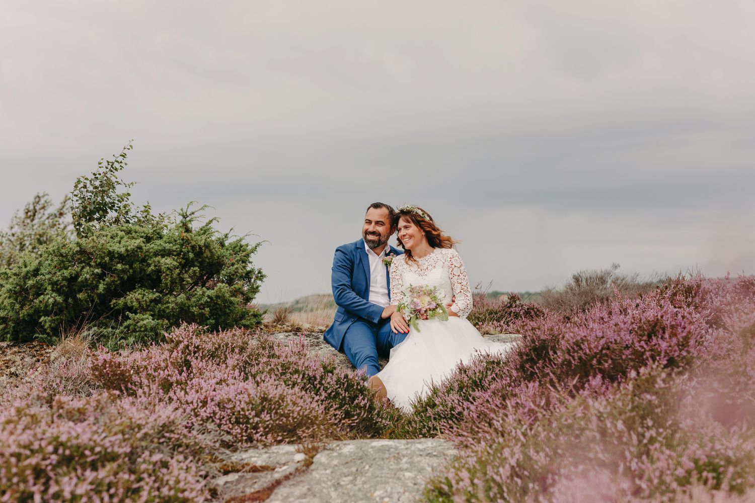 A couple sits together on a hillside surrounded by blooming purple heather under an overcast sky.