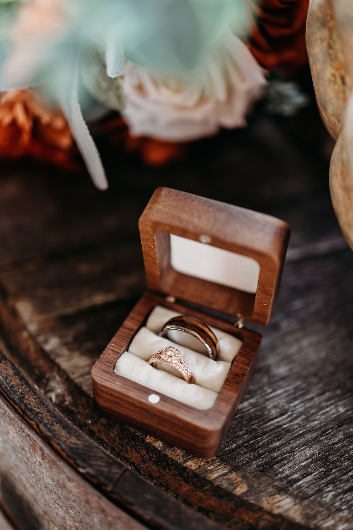 Two wedding rings displayed in an open wooden ring box on a rustic wood surface with floral bouquet in background.