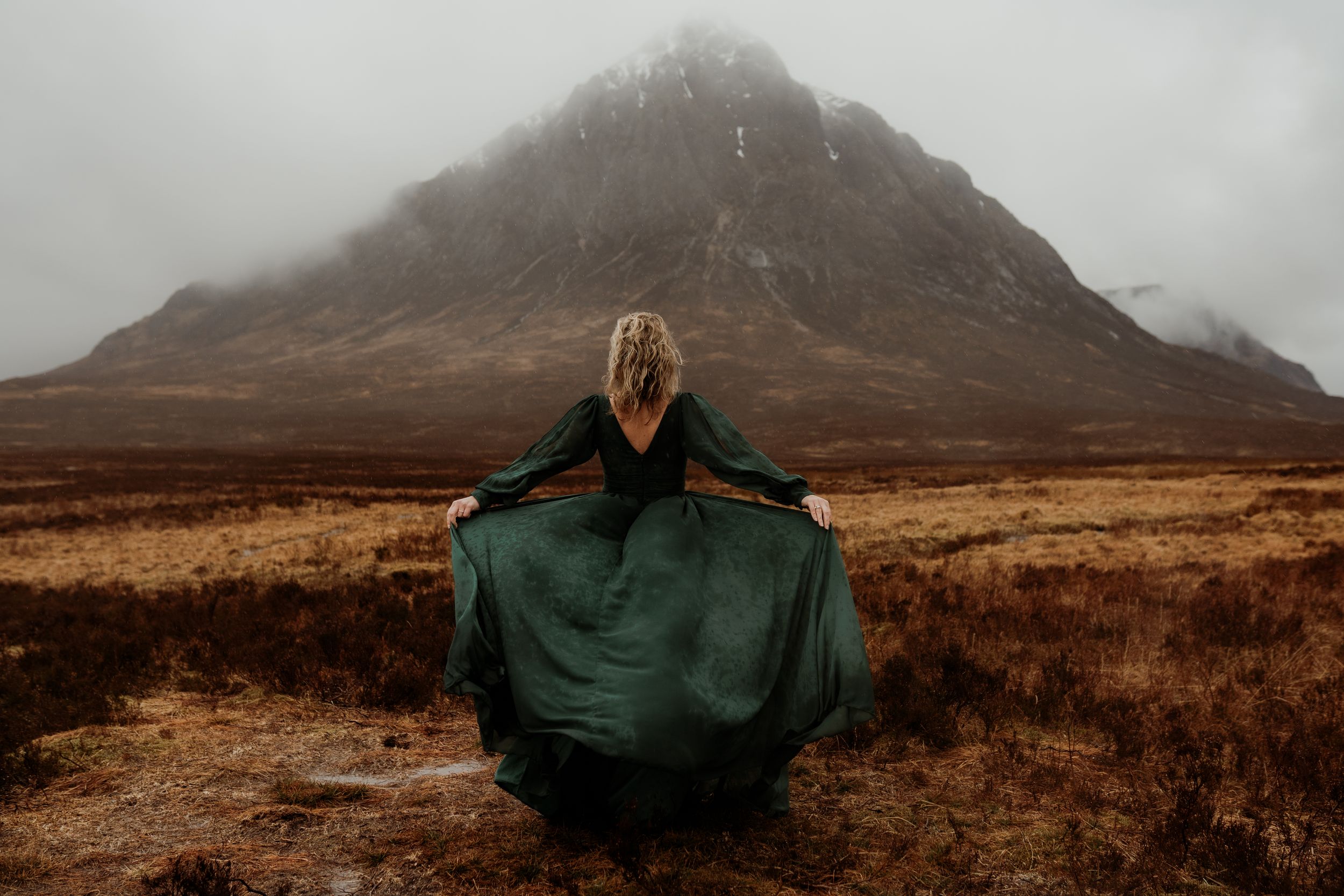 Woman in green dress stands before misty Scottish Highland mountain in dramatic foggy landscape.