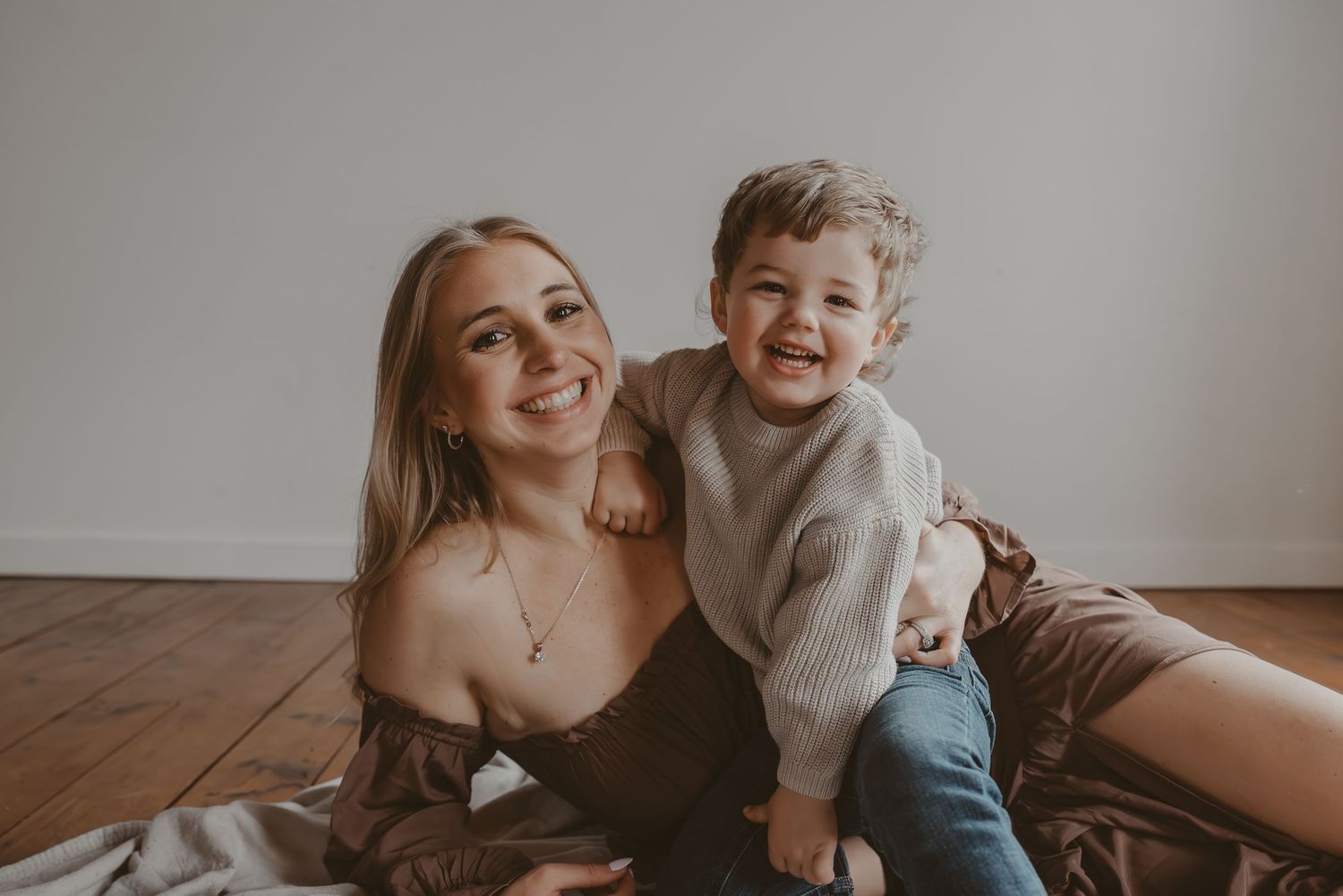 Woman in brown off-shoulder maxi dress holds hands with young child on white fabric on wooden floor.