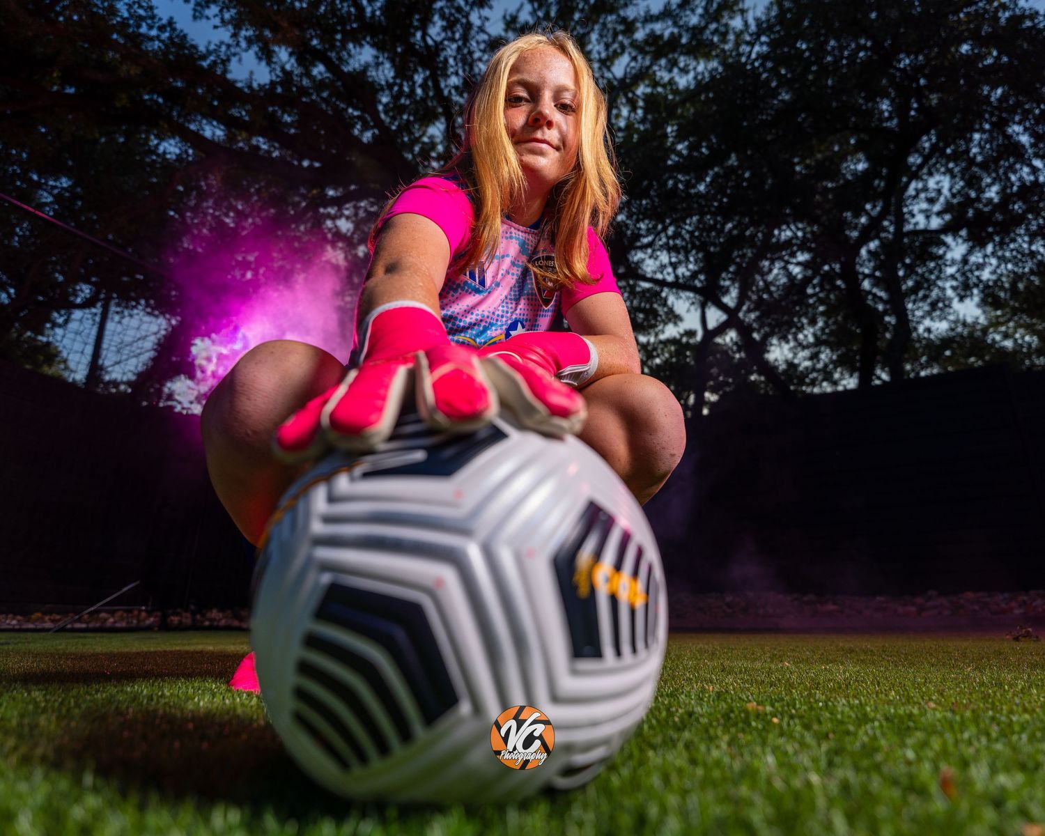 Female soccer player in pink gear crouches on grass field, touching a Nike soccer ball with dramatic lighting.
