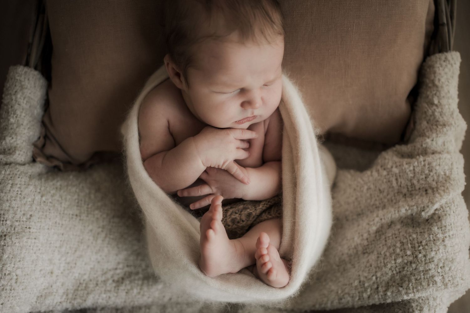 Newborn baby sleeping peacefully wrapped in a cream blanket inside a wooden bowl prop.