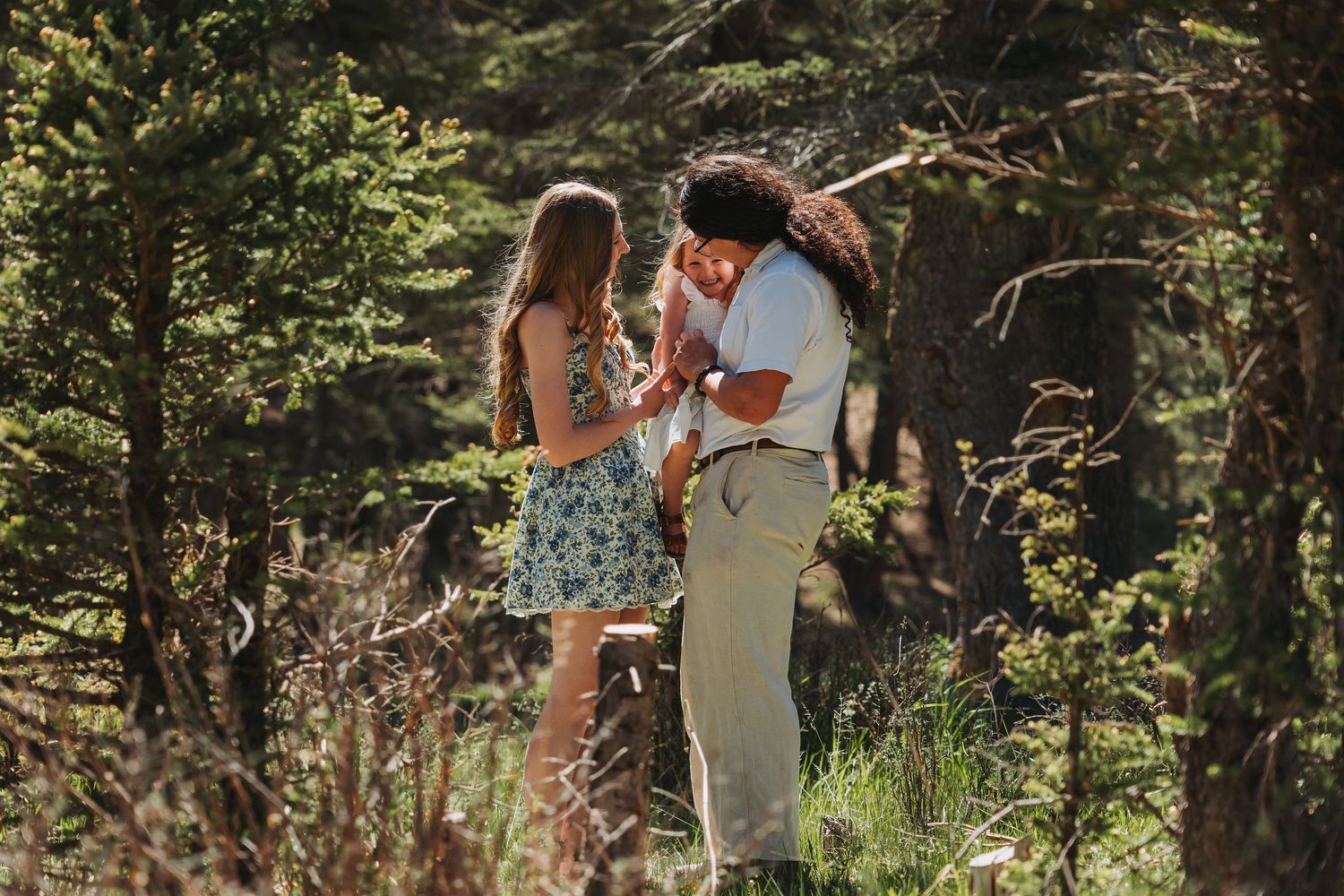 Couple sharing a romantic moment among tall trees in a sunlit forest, woman wearing floral dress and boots.