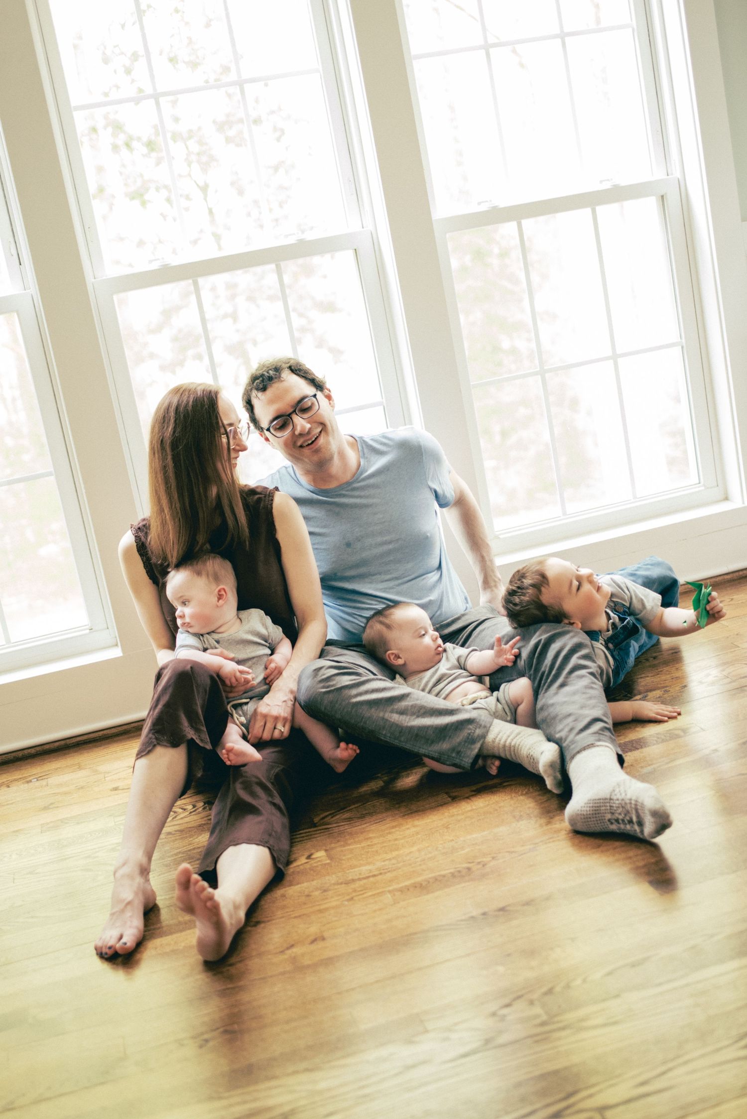 Happy family with two adults and three young children sitting together on hardwood floor by bright windows.