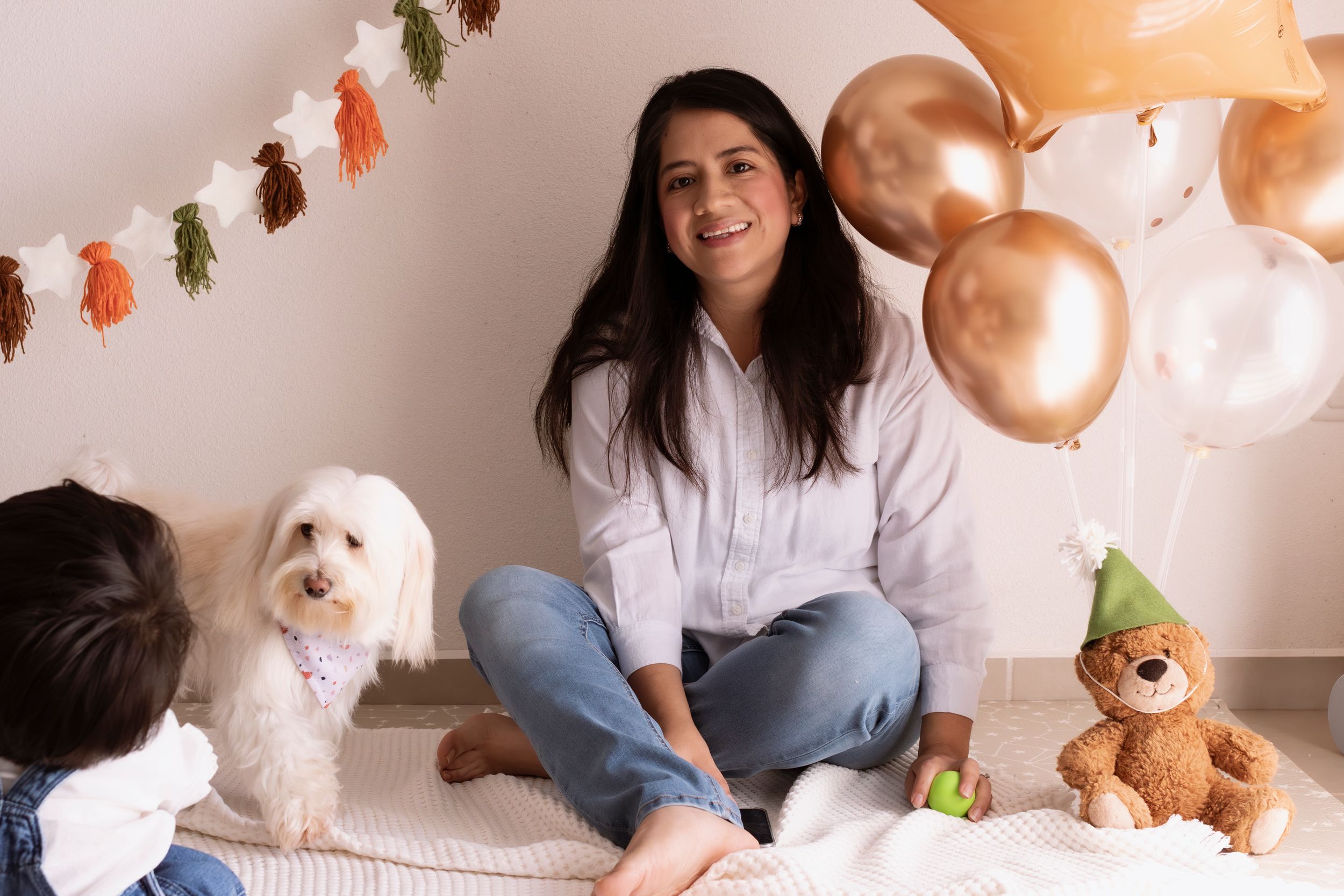 A cozy indoor scene with a white Maltese dog, decorative balloons, and a teddy bear during a celebration.