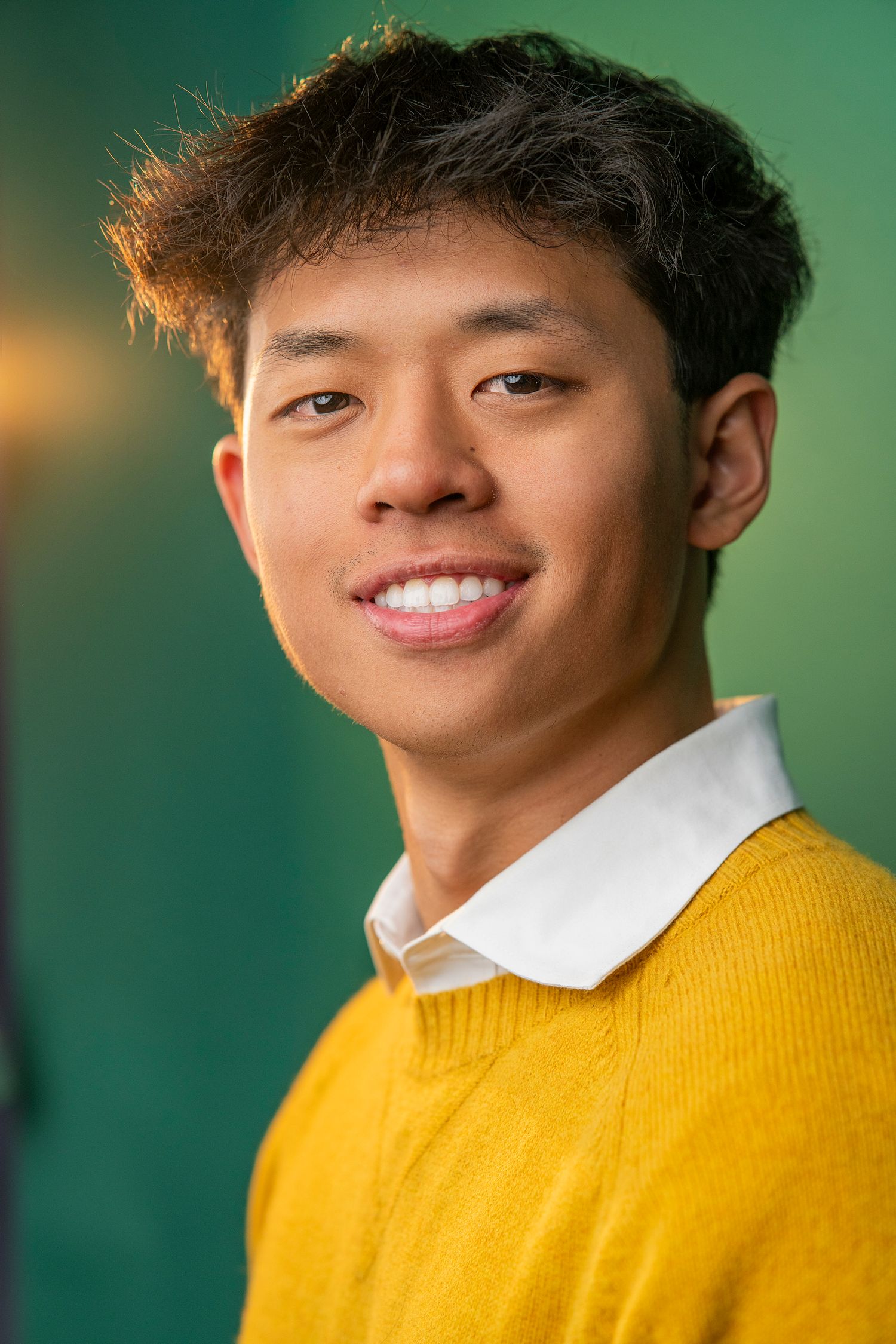 Smiling young Asian man wearing a yellow sweater over a white collared shirt against a green background.