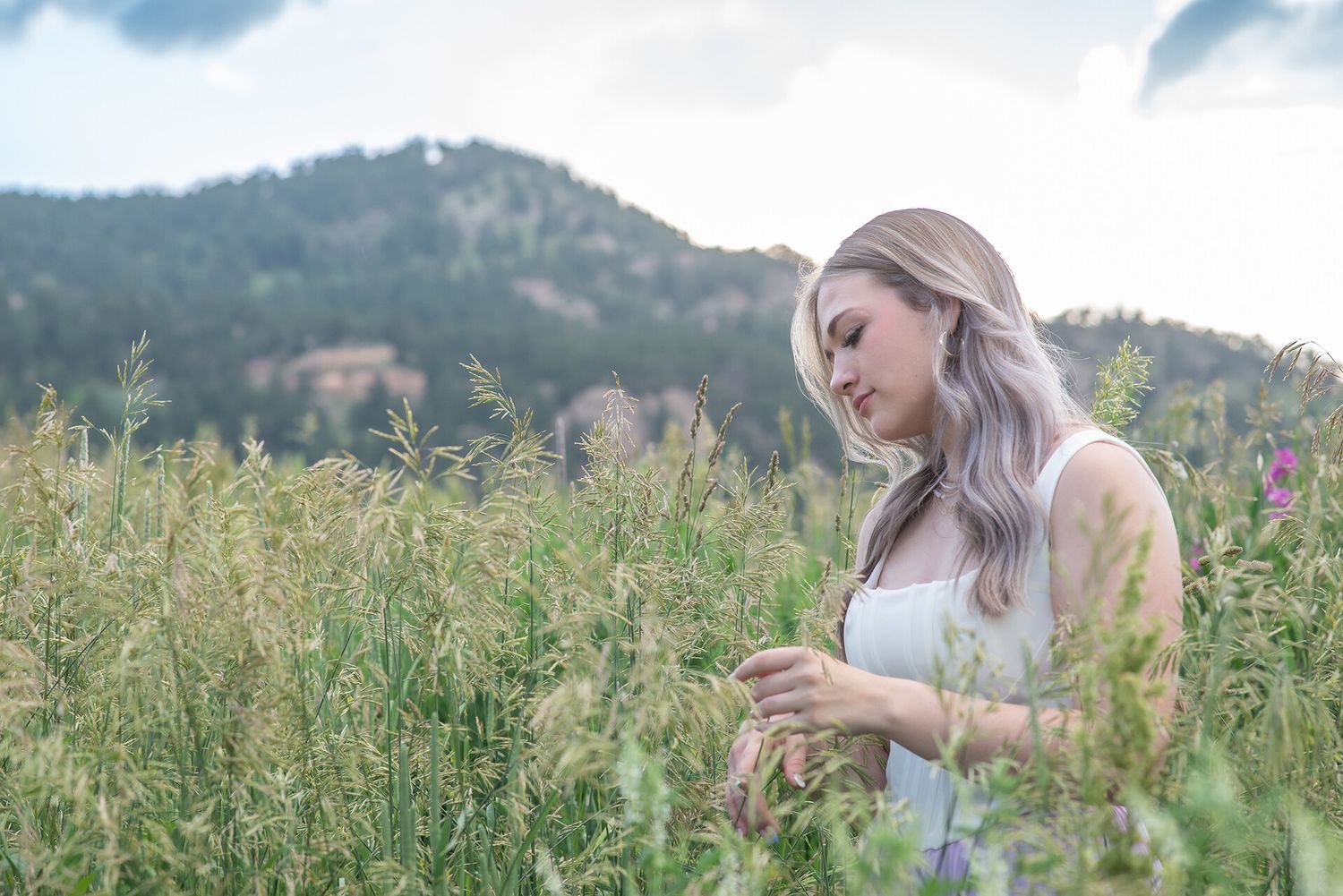 A young woman with blonde hair stands in a lush green field, gently touching wildflowers with mountains behind her.