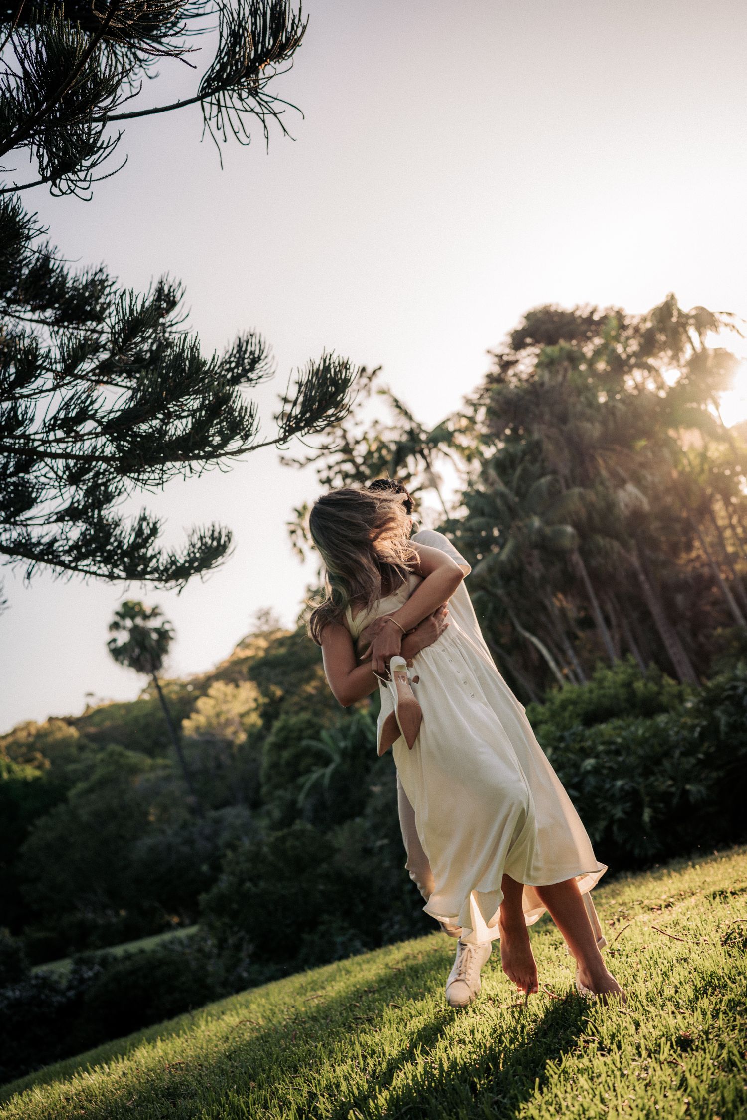 Woman in white dress twirling on green lawn at golden hour with lush tropical trees in background.
